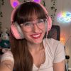 A selfie of a 30 year old white woman with long brain hair and bangs and green eyes. She is wearing a white t shirt, pink cat-ear headphones, and clear frame glasses. She is smiling at the camera.